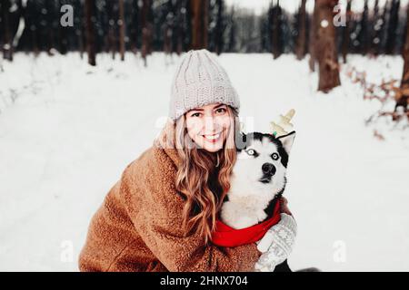 Giovane bella donna sorridente felice in pelliccia cappotto abbraccio cane Husky vestito di rosso sciarpa di Natale e corna di cervo festive. Ritratto foto di ragazza e fare Foto Stock