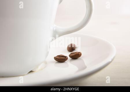 Tazza bianca in porcellana con caffè su piastra bianca con fagioli su tavola bianca in legno alla vista macro della colazione. Vista frontale. Composizione orizzontale. Foto Stock