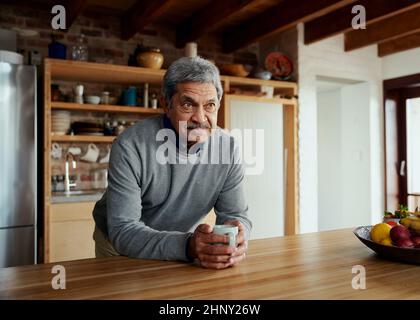 Pensionato maschile anziano e biraciale pensionato che si appoggia sul moderno banco della cucina, tenendo in mano il caffè del mattino. Foto Stock