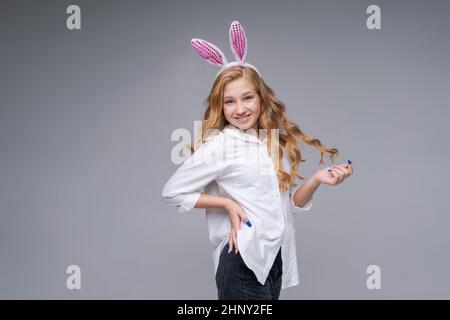 Giovane bella donna in carino pasqua coniglietto orecchie fa un gesto normale, gioca con la sua mano con i capelli lunghi, in una camicia bianca su uno sfondo grigio sorride felicemente. Ragazza caucasica in studio Foto Stock