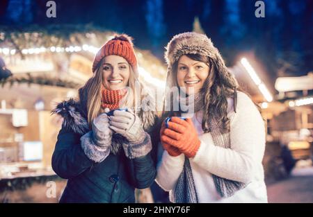 Due donne con VIN brulé caldo in tazze di fronte al mercatino di Natale stand Foto Stock