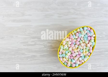 Uovo di Pasqua decorativo e mini marshmallows multicolore su sfondo bianco in legno con spazio copia per congratulazioni e desideri Foto Stock