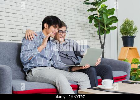 Uomo d'affari asiatico senior con computer portatile discutere insieme con il team giovane in ufficio. Padre uomo e suo figlio si siedono sul divano a parlare in video Foto Stock
