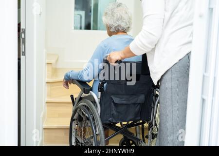 Aiuto dell'operatore sanitario asiatico o anziano anziana donna seduta sedia a rotelle sostenere le scale in casa. Foto Stock