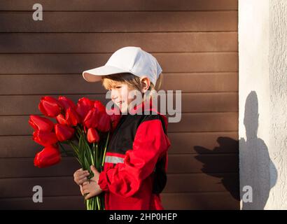 ritratto del profilo di un gioioso ragazzo di 5 anni in uniforme rossa, berretto e con un bouquet di tulipani rossi. Concetto di consegna, corriere piccolo, fiori con lov Foto Stock