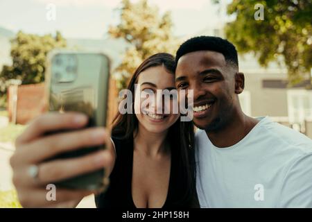 coppia biraciale sorridente mentre si prendono selfie nel parco Foto Stock