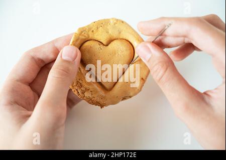 Mani closeup graffio marrone zucchero caramello caramelle biscotti con un ago di metallo a forma di cuore su uno sfondo bianco Foto Stock