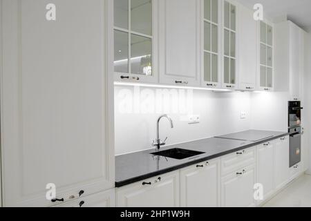Vista laterale moderna ed elegante cucina con cassetti e armadi bianchi, piano in marmo nero e lavandino incorporato con rubinetto d'argento Foto Stock