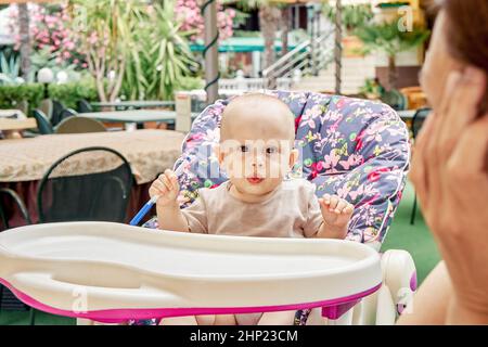 Il bambino siede nella poltrona del ristorante all'aperto sotto la cura della madre. Il bambino gioca con la penna blu contro le piante colorate e fiorenti del giardino Foto Stock