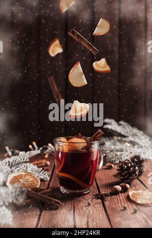 Bicchiere di Natale o tazza con VIN brulé su tavola di legno e sfondo con bastone di cannella levitante, arancio secco o sublimato, fette di mela, a Foto Stock