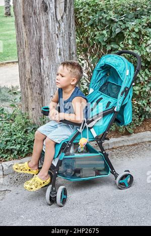 Il preschooler frena seduto in un passeggino blu di bambino fratello che cammina nel parco. Il ragazzo dai capelli discrete siede e si stanca dalla lunga passeggiata durante le vacanze estive Foto Stock