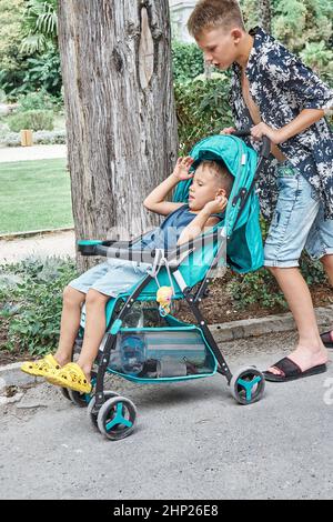 Il ragazzo di scuola biondo spinge il passeggino con il fratello più giovane seduto nel parco. Il ragazzo dai capelli discrete riposa e si schiena stanco dalla lunga passeggiata in estate Foto Stock