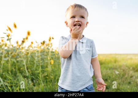 Ragazzino che gioca sullo sfondo della campagna e sorride. Tre anni di divertimento in campo di girasoli al tramonto. Stile di vita estivo all'aperto Foto Stock