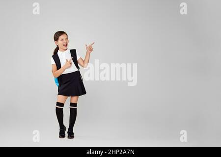 eccitata piccola studentessa carina con zaino che punta allo spazio di copia su sfondo grigio. Foto Stock