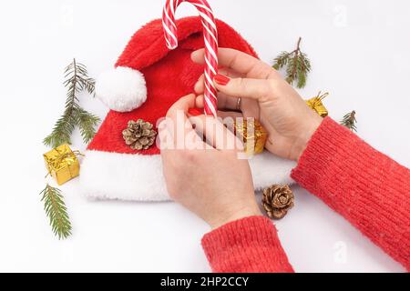 Mani femminili che tengono un lecca-lecca. Ramo di abete, coni, canna di caramello, regalo, nel cappello di Babbo Natale su sfondo bianco. Isolato Foto Stock