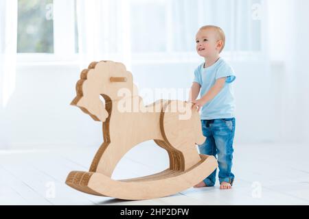 Carino, sorridente, bianco, ragazzo di due anni in t-shirt blu e jeans dondolanti su cavallo di legno fatto a mano. Il bambino si diverte con il giocattolo dei pony. Concetto di Foto Stock