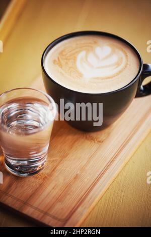 Cappuccino caldo in una tazza, posto su un vassoio di legno accanto a un bicchiere d'acqua limpida nel bar retrò. Foto Stock