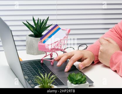 donna in un maglione rosa effettua acquisti online con un laptop, una carta di credito in mano. Donna seduta ad un tavolo bianco Foto Stock