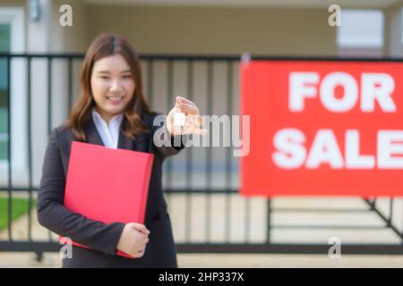 Agente immobiliare asiatico o donna immobiliare sorridente e tenendo file rosso con mostrare la chiave di casa di fronte alla casa. Foto Stock
