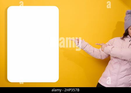 donna in una giacca rosa invernale e cappello mostra con le mani a lato su un cartellone bianco bianco bianco su sfondo giallo. Luogo per gli annunci, adv Foto Stock