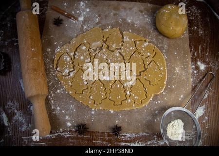 Nello strato di pasta arrotolato, molti alberi di Natale sono tagliati, che sono contrassegnati con contorni, l'impasto si trova su forno pergamena, accanto ad esso giace Foto Stock