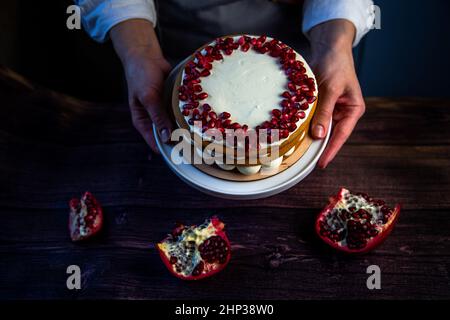 Un dolce con pan di Spagna, decorato con crema bianca e semi di melograno sulla sommità, è tenuto in cerchio da uno chef pasticceria su sfondo di w scuro Foto Stock