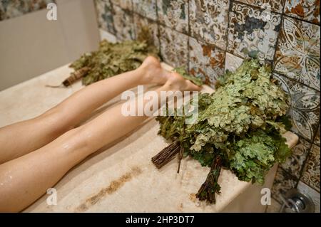 Vista dall'alto gamba femmina su panca in legno. Riposo donna in sauna spa o bagno russo con scopa di quercia Foto Stock