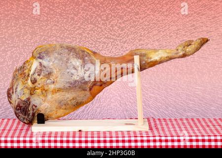 Cucina spagnola. Primo piano di una gamba anteriore di un prosciutto spagnolo Serrano su un piedistallo di legno su una tovaglia a scacchi rossa sopra astratto cubo rosa blocco raster bac Foto Stock