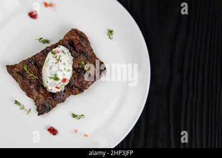 Bistecca di carne di manzo alla griglia su un piatto con salsa di verdure bianche decorata da erbe e bacche. Sfondo di legno scuro Foto Stock