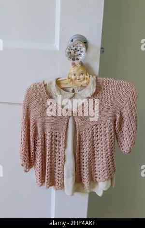 Maglione vintage rosa con crocchette appeso sulla blusa ricamata vintage bianca su un antico gancio in plastica con testa delle bambine sulla porta in vetro tagliata k Foto Stock