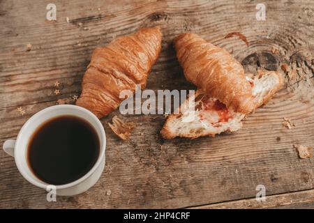 tagliare il croissant con burro e marmellata di fragole con una tazza di caffè, su sfondo di legno, vista dall'alto. Foto Stock