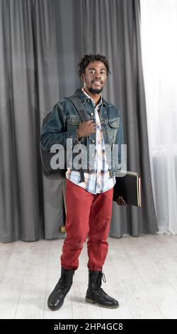 Giovane uomo afro-americano alla moda con barba che tiene libri e zaino con giacca in denim pronta per andare a studi. Bel giovane studente sulla strada per la scuola. Concetto di scuola. Concetto di studio. Foto Stock