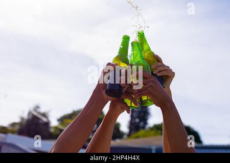 Mani di amici biraciali maschi e femmine che tostano bottiglie di birra contro il cielo Foto Stock