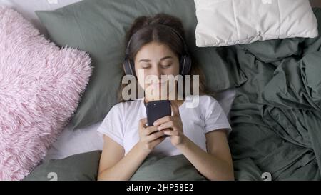 Happy Young Woman in Headphones ascolta musica mentre si trova in un letto accogliente Foto Stock
