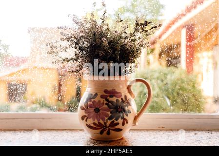 Vaso vecchio con fiori secchi in una finestra in un giorno di pioggia d'autunno nostalgico, film grano aggiunto. Foto Stock