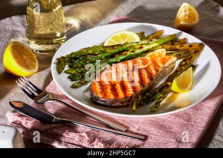 bistecca di salmone alla griglia con asparagi arrosto e limone su un piatto bianco su una tovaglia di lino con un decanter di vino bianco Foto Stock