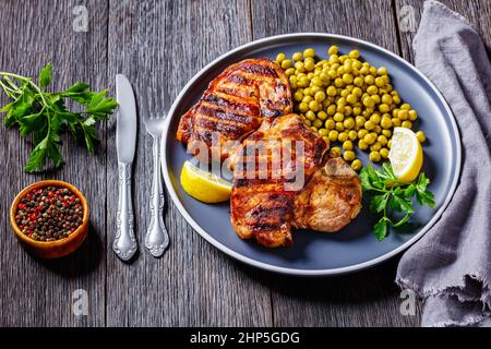 bistecche di carne di maiale alla griglia con piselli verdi bolliti e limone su un tavolo di legno con forchetta e coltello Foto Stock