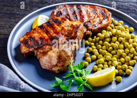 bistecche di carne di maiale alla griglia con piselli verdi bolliti e limone su un piatto su un tavolo di legno, vista orizzontale dall'alto, primo piano Foto Stock