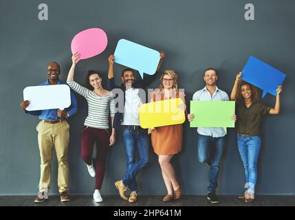 Lasciate che vi aiutiamo a far uscire la vostra parola. Studio girato di un gruppo diversificato di persone che tengono in su bolle di discorso su uno sfondo grigio. Foto Stock