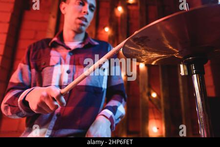 Ritagliare un musicista maschile di talento in camicia a scacchi che suona tamburi con bastoncini di batteria mentre si esibisce la canzone sul palco con lampade incandescenti durante il concerto Foto Stock