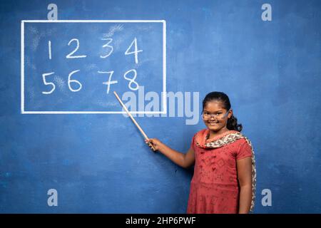 Bambina con bastone in mano che punta a bordo agendo come insegnante o professore - concetto di sogni d'infanzia, istruzione e stile di vita giocoso Foto Stock