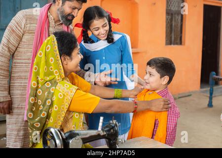 La madre indiana controlla le dimensioni dei panni su suo figlio mentre marito e figlia lo guarda, cucendo i panni sulla macchina da cucire. Piccola famiglia, stile di vita. Foto Stock