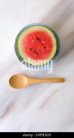 Posare piatto di un piccolo cocomero rotondo tagliato a metà, e un cucchiaio di legno accanto ad esso. Posizionato su un bancone in marmo. Foto Stock