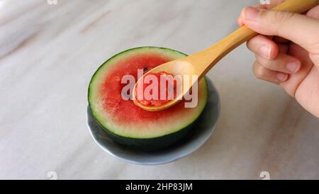 Mano che tiene un cucchiaio di legno, scavando in un piccolo cocomero tagliato a metà. Foto Stock