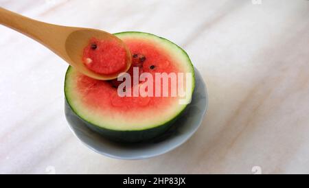 Cucchiaio di legno che raccoglie una parte di un piccolo cocomero tagliato a metà. Con sfondo in marmo. Foto Stock