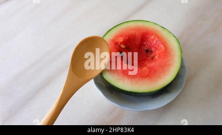 Piccolo cocomero tagliato a metà, con un piccolo buco al centro, e un cucchiaio di legno accanto ad esso. Su una superficie in marmo. Foto Stock
