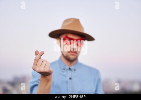 Uomo caucasico con maschera rossa supereroe e camicia in jeans che si posa davanti alla fotocamera mostrando il cuore coreano o il gesto 'cuore mini' all'aperto al tramonto con vista urbana. Immagine di alta qualità Foto Stock