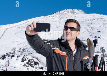 Bel sciatore che prende un selfie su una montagna innevata mentre tiene i suoi sci Foto Stock