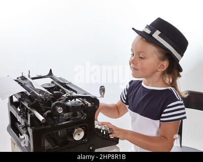 Ragazza piccola graziosa nel cappello che batte sulla macchina da scrivere antica su sfondo bianco. Concetto di istruzione.fcool Foto Stock