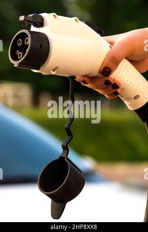 Una mano femmina prende il cavo di carica di un veicolo elettrico. Il conducente con un cavo per la ricarica di un'auto elettrica sullo sfondo di un'auto. Verde Foto Stock